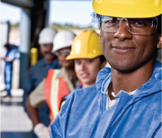 Young man wearing a hard hat standing in front of a line of men and women, also wearing hard hats.