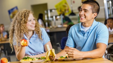 A teen boy and girl wearing school unifroms and laughing over lunch.  The girl is holding an apple and the boy is holding a banana.  