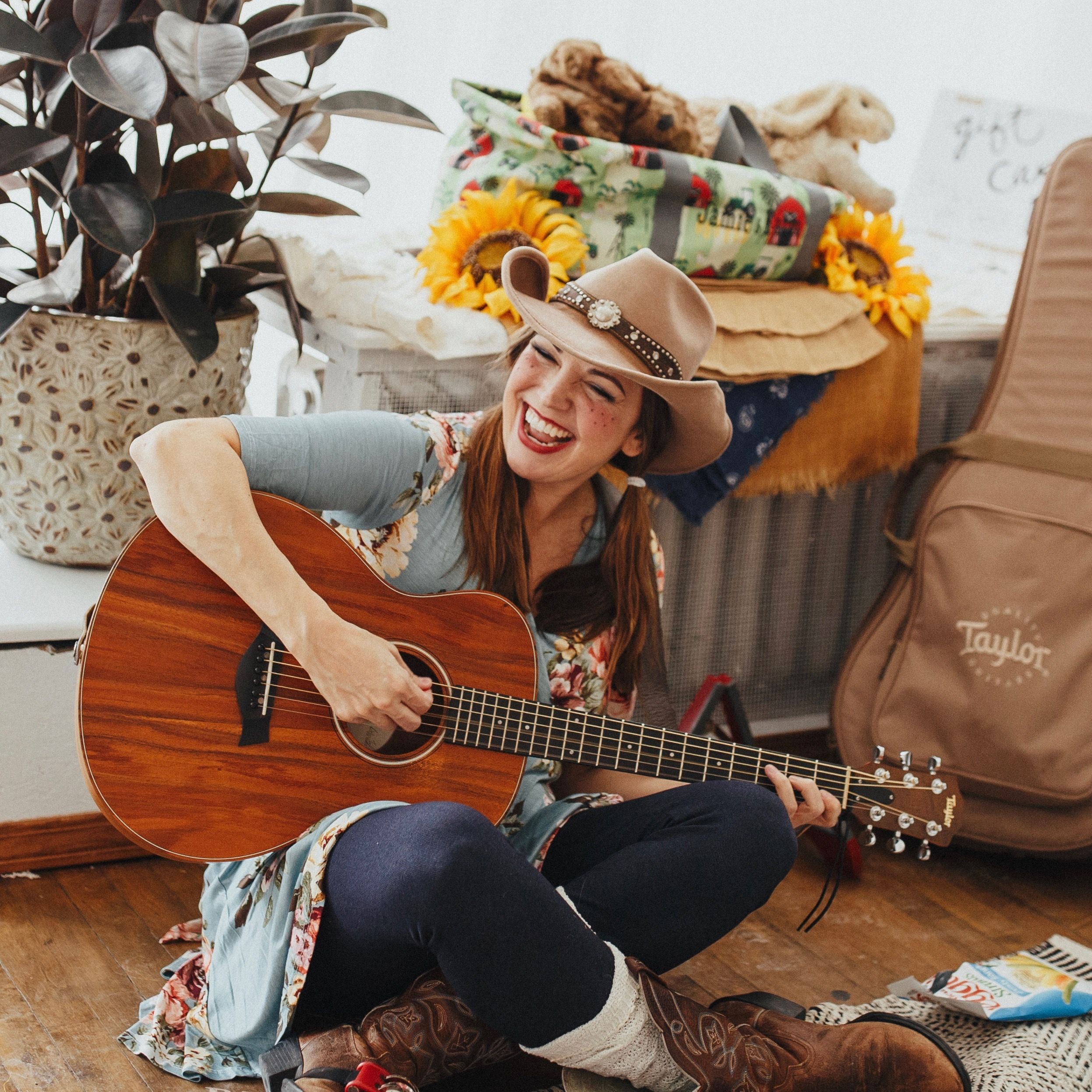 Woman in a cowboy hat, sitting on the wood floor, playing the guitar.