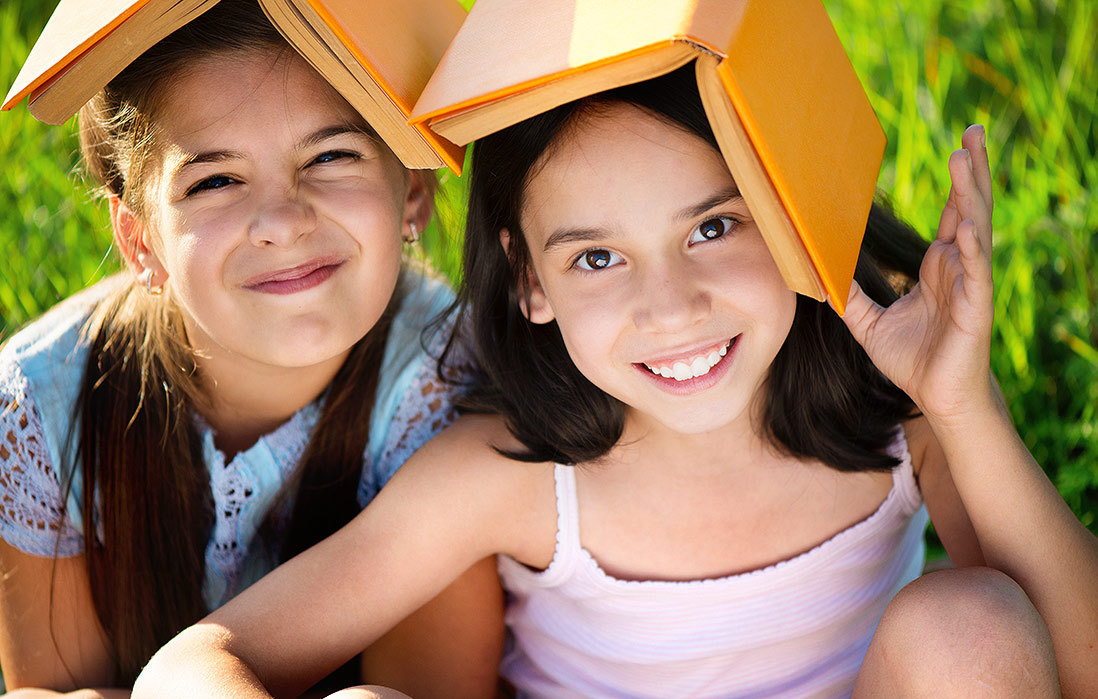 Two girls, outdoors, wil open books on their heads.