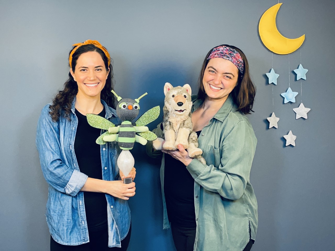 Two women, each holding a puppet.  A paper moon hangs in the background.