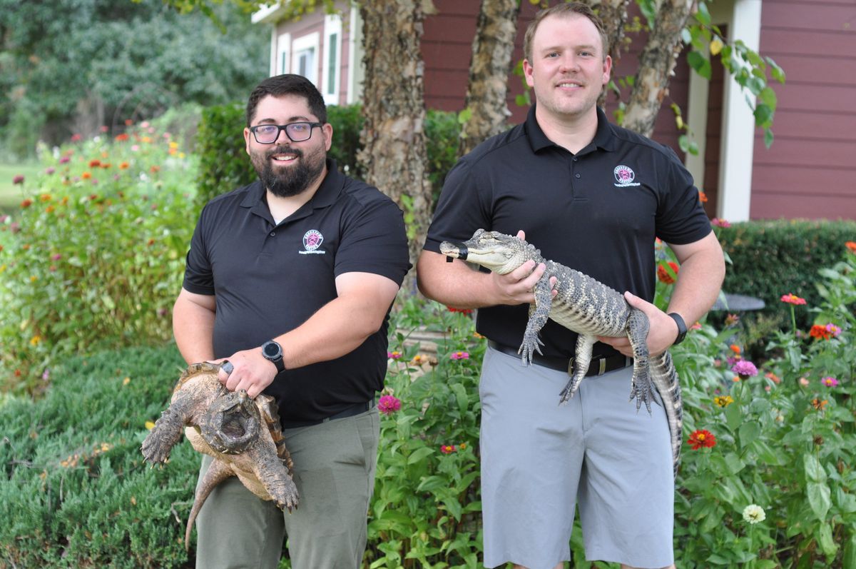 Two adult men in matching shirts, one with a beard, holding a large turtle and a small alligator.