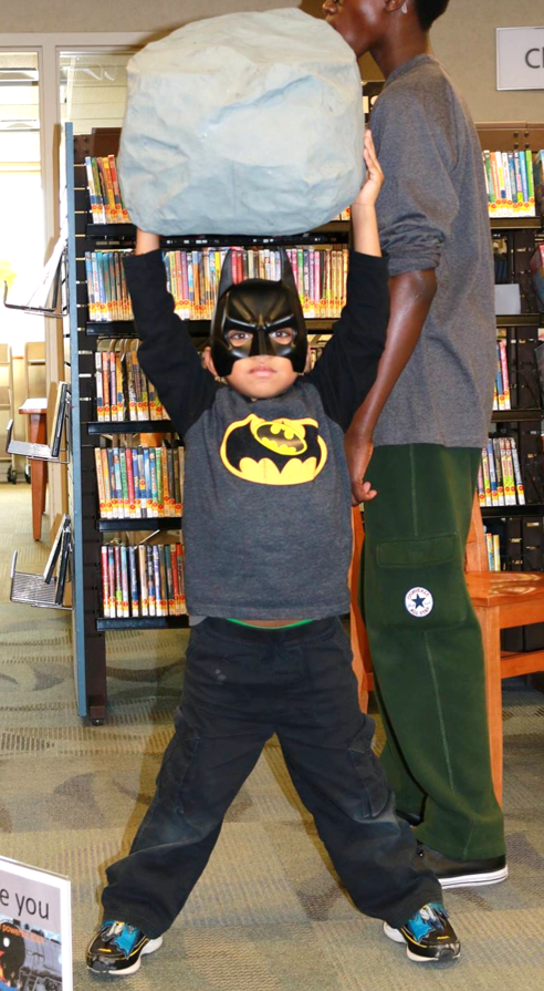 A young boy dressed up as Batman, holding a large papier mache rock over his head.