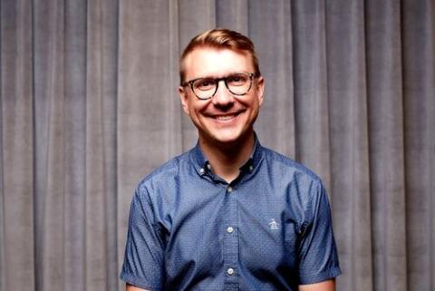 A bespectacled man smiling directly into the camera, wearing a button-down, short-sleeved shirt.