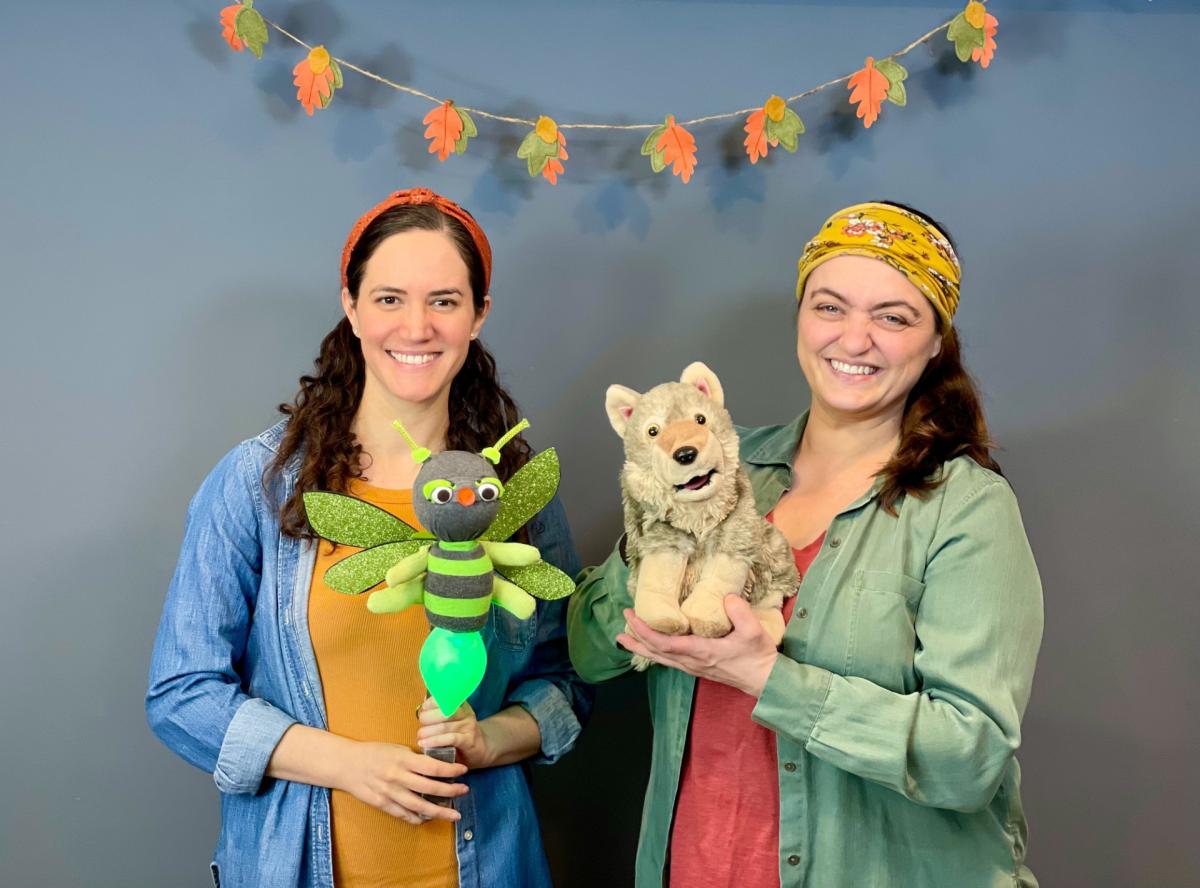 Two women holding puppets in front of a leaf garland.